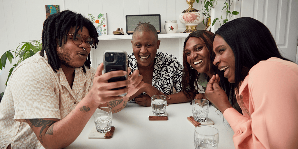 Group of four friends laughing together while looking at a phone at a table