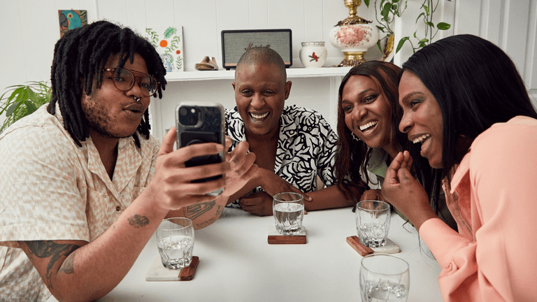 Group of four friends laughing together while looking at a phone at a table