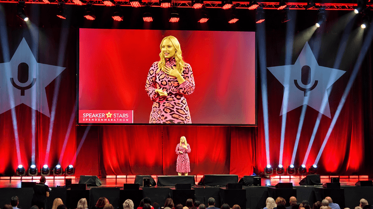 A speaker in a pink dress presents on stage at a conference with an audience.