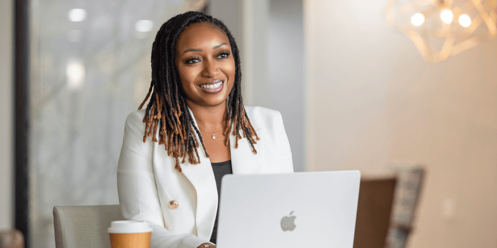 Smiling woman with dreadlocks sitting at a table with a laptop and coffee cup
