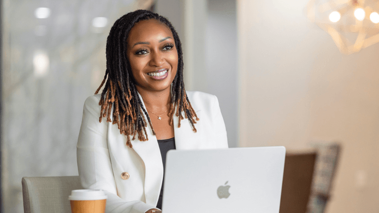 Smiling woman with dreadlocks sitting at a table with a laptop and coffee cup