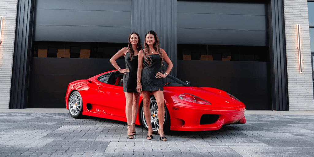 Two women pose in front of a red sports car outside a modern building
