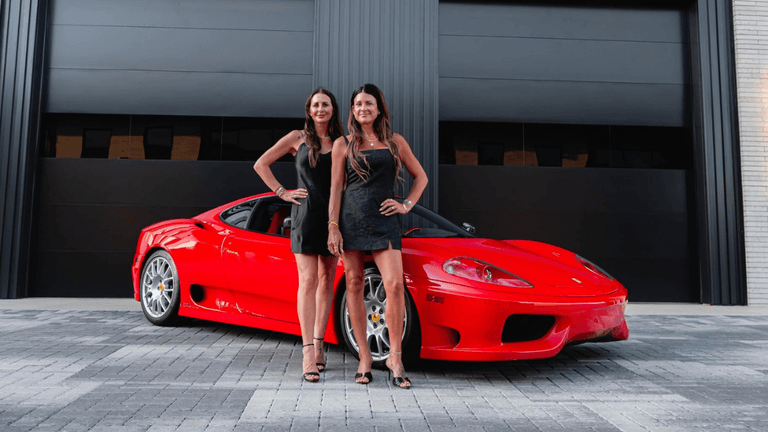 Two women pose in front of a red sports car outside a modern building