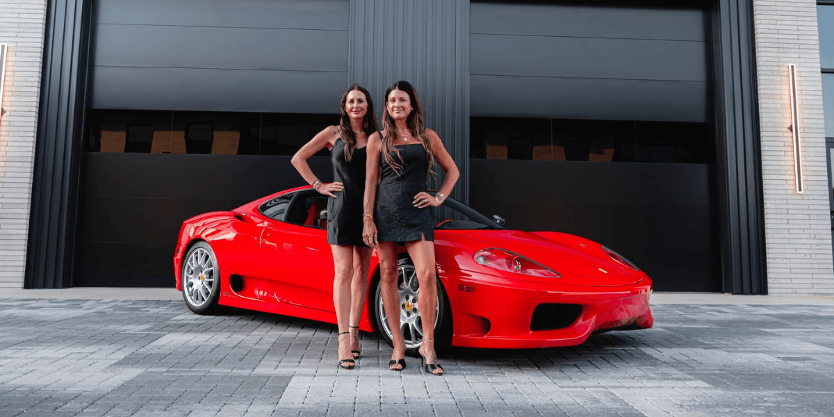 Two women pose in front of a red sports car outside a modern building