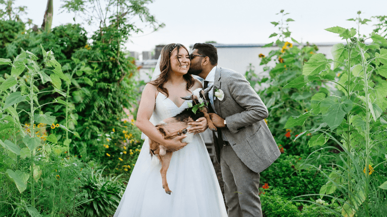 A couple in wedding attire holding a dog, surrounded by a lush garden