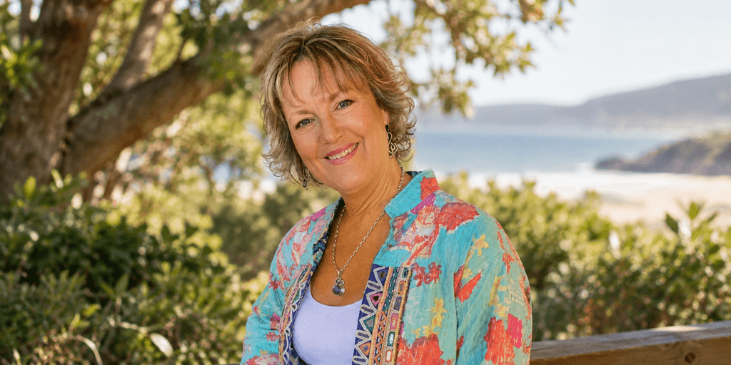 Smiling woman in colorful attire sitting outdoors with a scenic background of water and greenery