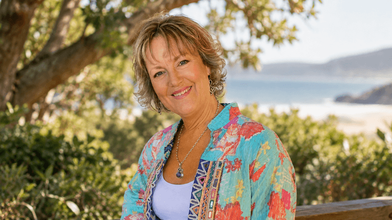 Smiling woman in colorful attire sitting outdoors with a scenic background of water and greenery