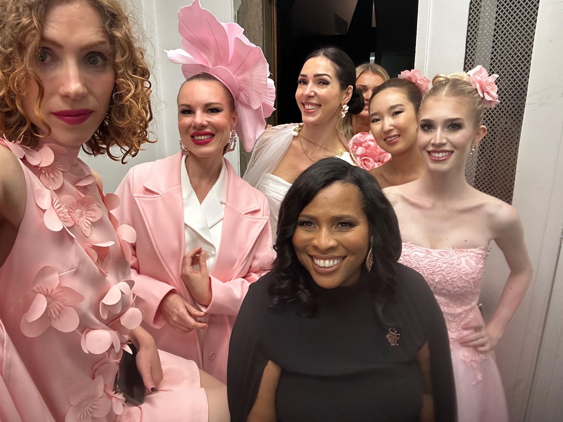 Six women in elegant dresses and floral accessories pose together backstage at an event