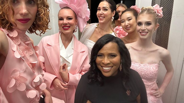 Six women in elegant dresses and floral accessories pose together backstage at an event