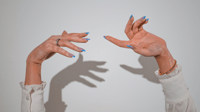Two hands with stylish blue nails reaching outward, casting shadows against a white background