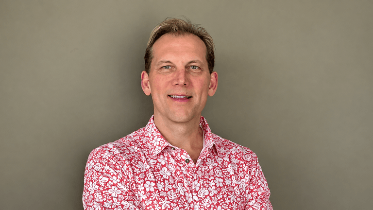 Smiling man in a floral shirt posing with arms crossed against a neutral background