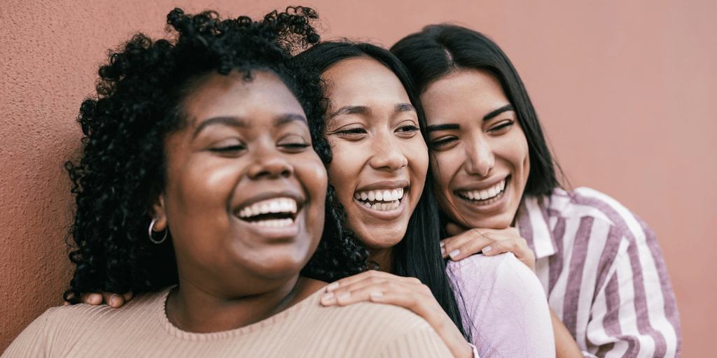 Three women smiling and embracing against a reddish wall background