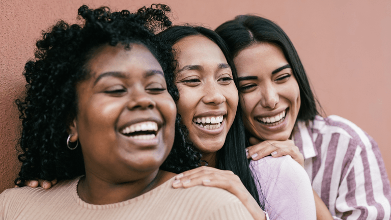 Three women smiling and embracing against a reddish wall background