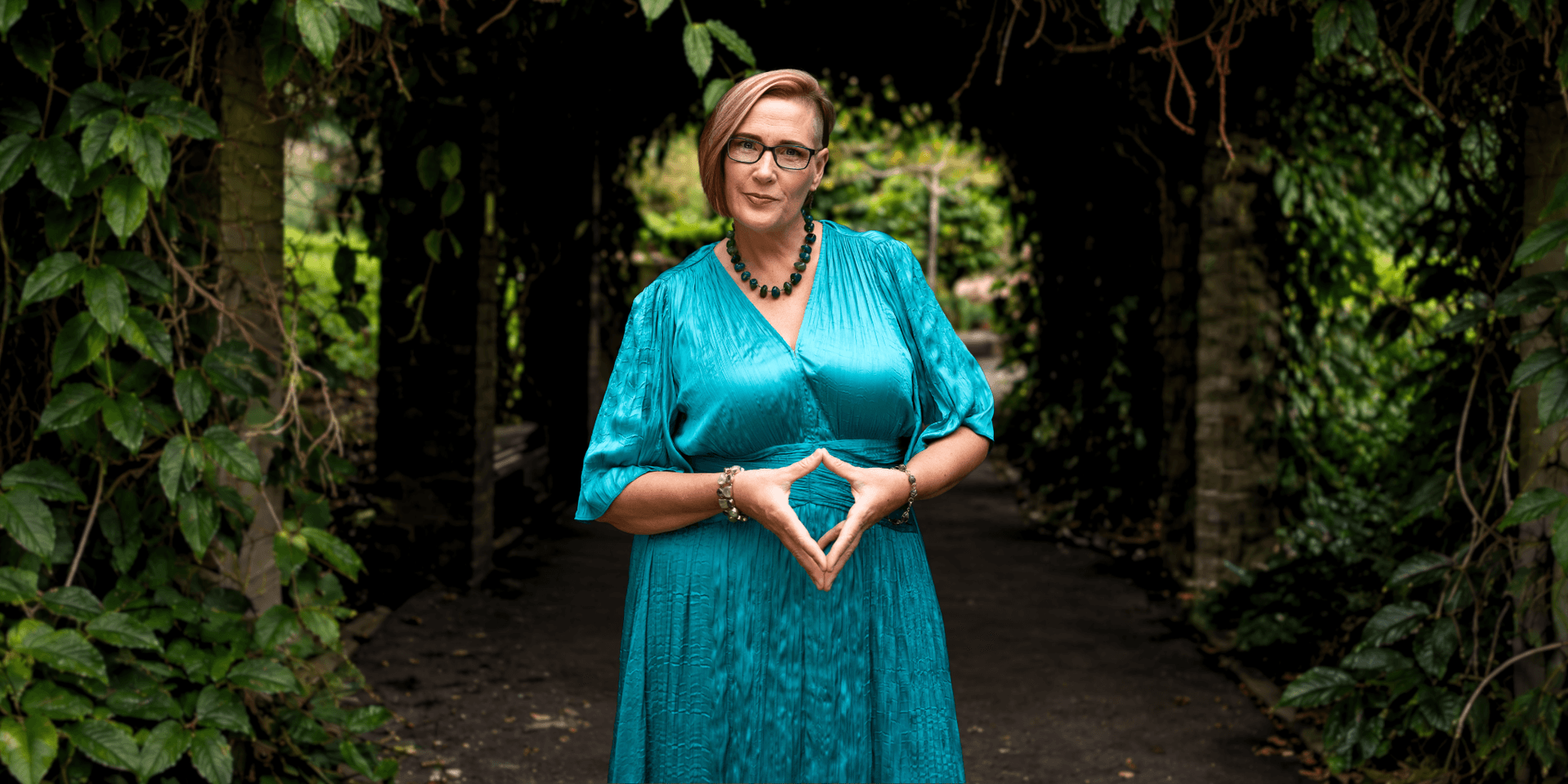 A woman in a teal dress stands confidently in a green garden archway