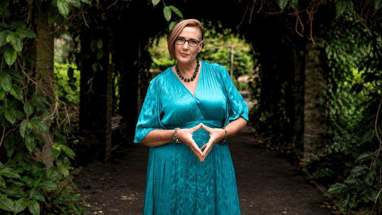 A woman in a teal dress stands confidently in a green garden archway