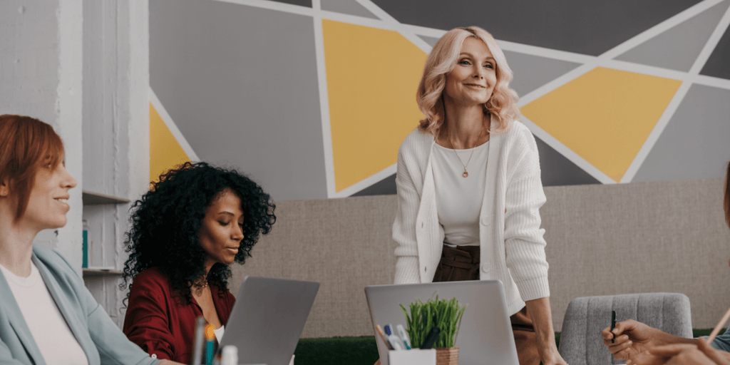 Four women in a modern workspace with laptops, engaged in a discussion.