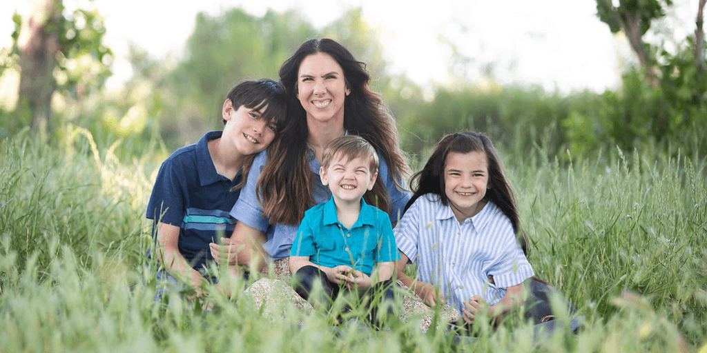 A mother sits in a grassy field with three smiling children.
