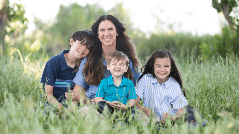 A mother sits in a grassy field with three smiling children.