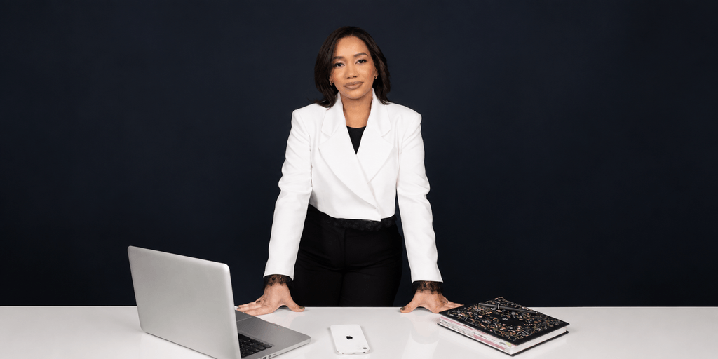 Woman in a white blazer stands at a desk with a laptop and notebooks