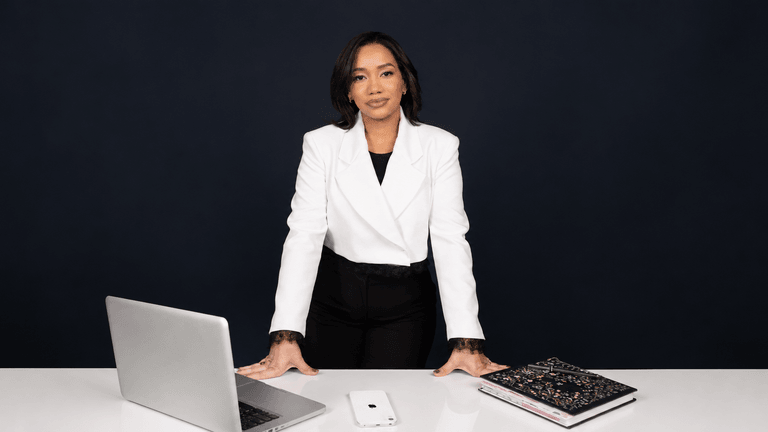 Woman in a white blazer stands at a desk with a laptop and notebooks