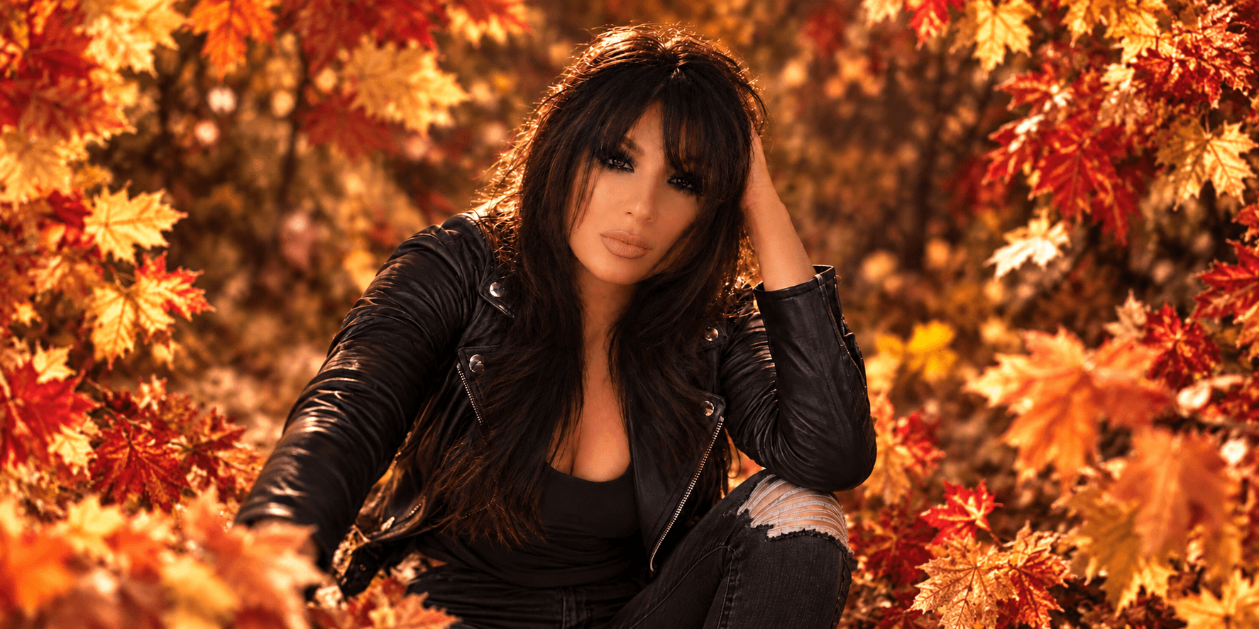 A woman in a black leather jacket sits among vibrant autumn leaves.