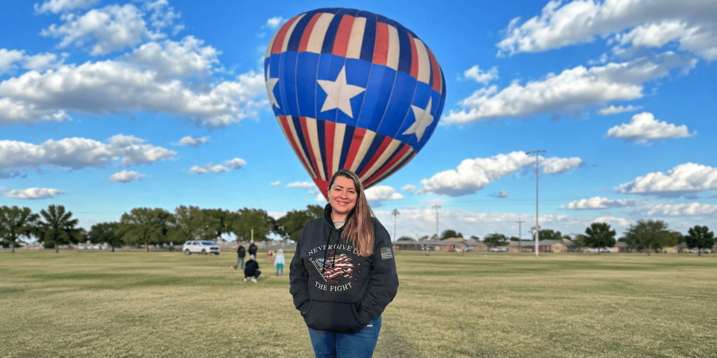 Person in a hoodie standing in a grassy field with a hot air balloon above