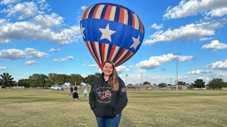 Person in a hoodie standing in a grassy field with a hot air balloon above