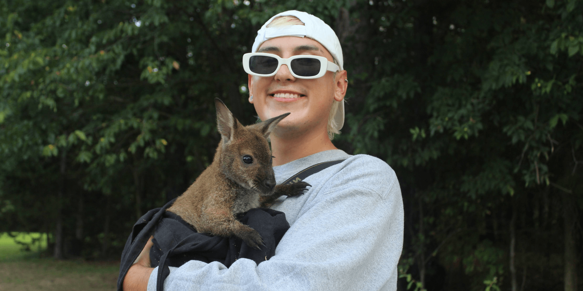 A smiling person holds a small wallaby against their chest in a natural setting