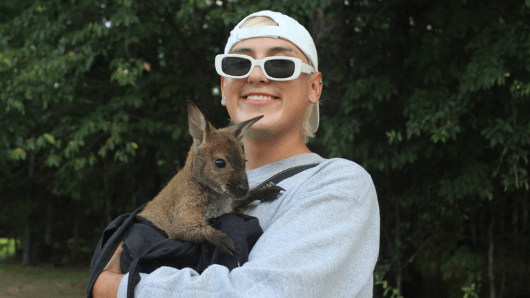 A smiling person holds a small wallaby against their chest in a natural setting