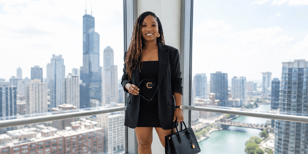 A woman in a black blazer and dress stands by a window with city views