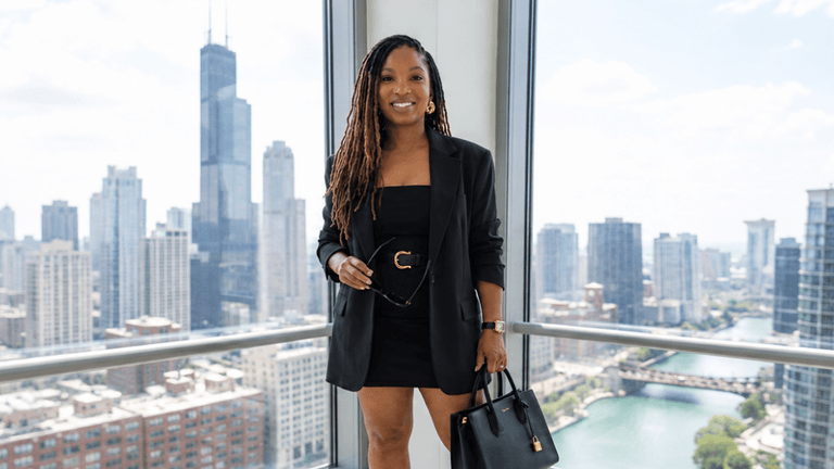 A woman in a black blazer and dress stands by a window with city views