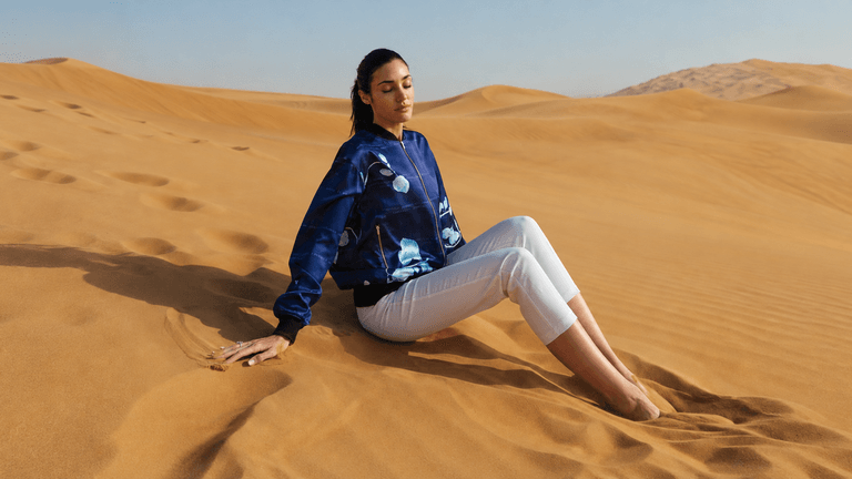 Model sitting in desert sand wearing a blue jacket and white pants