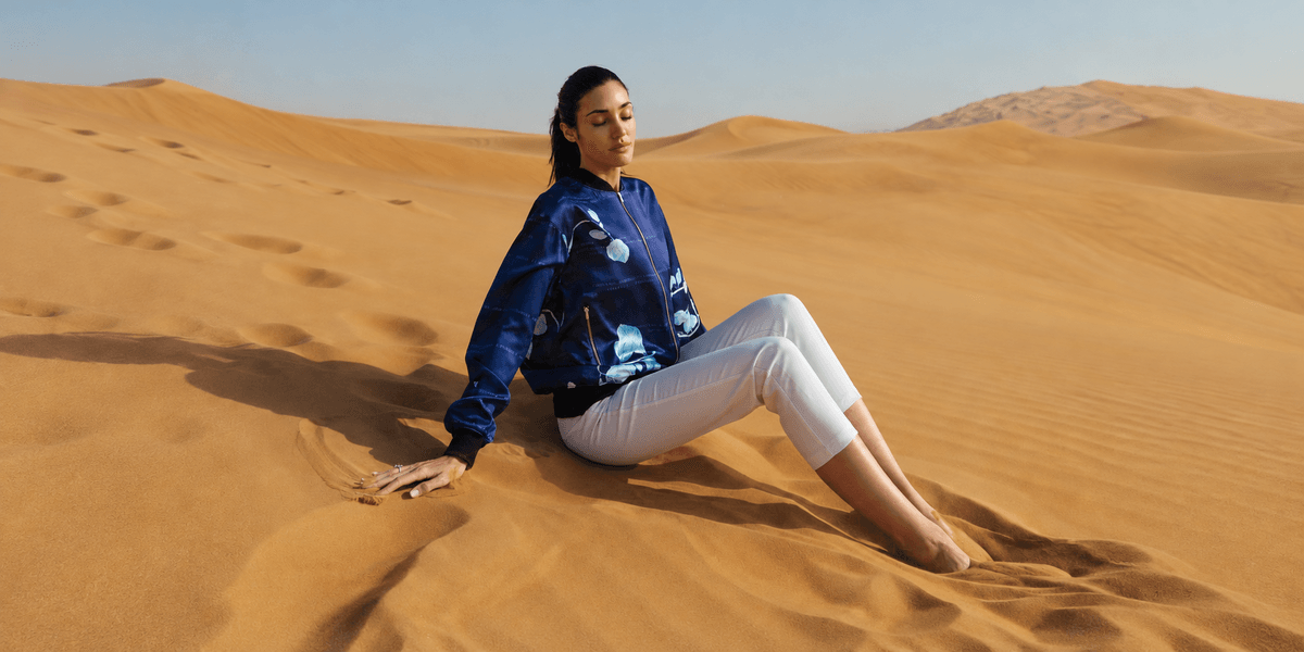 Model sitting in desert sand wearing a blue jacket and white pants