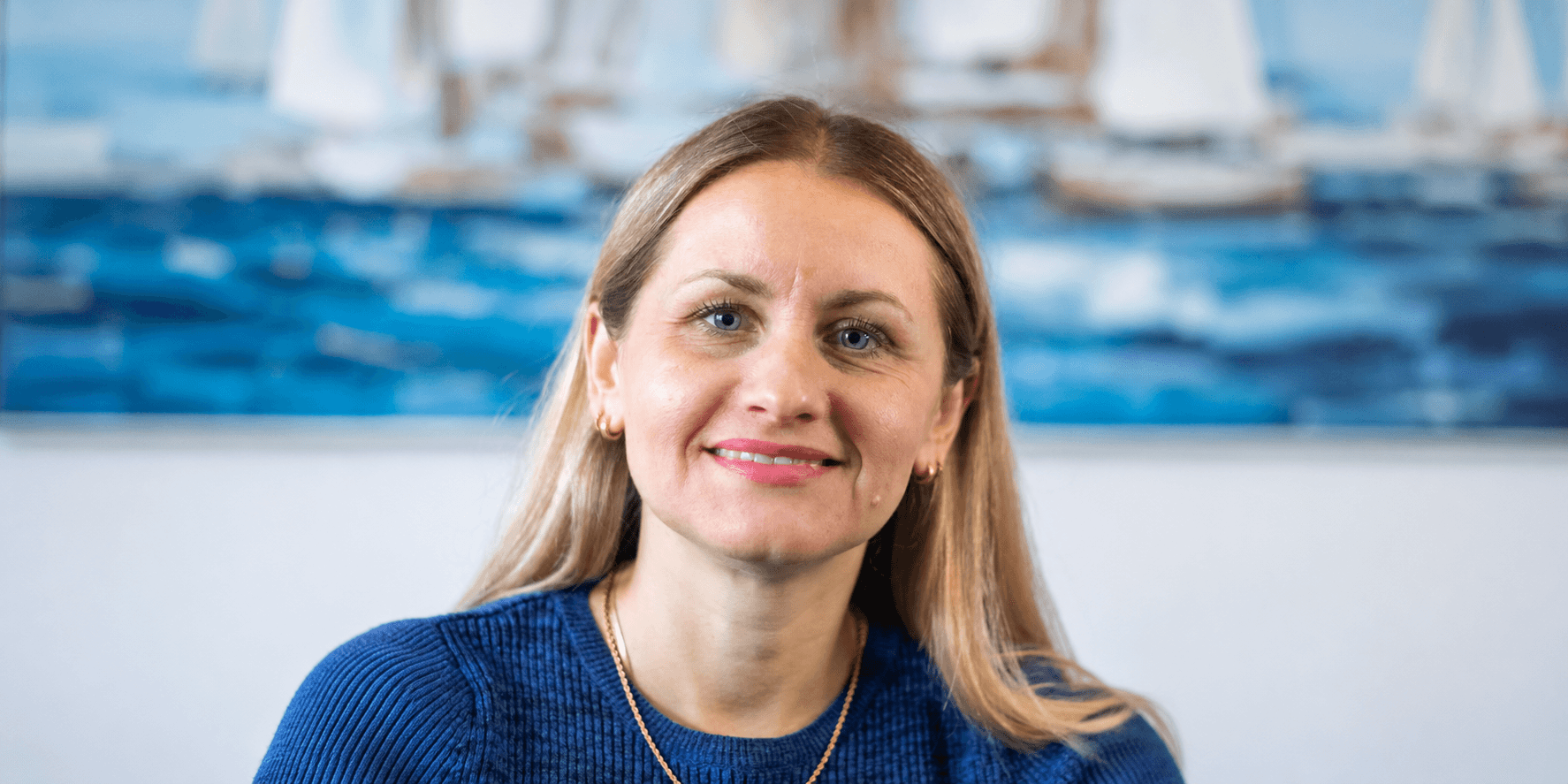 A woman with long blonde hair, smiling, wearing a blue sweater against a nautical backdrop