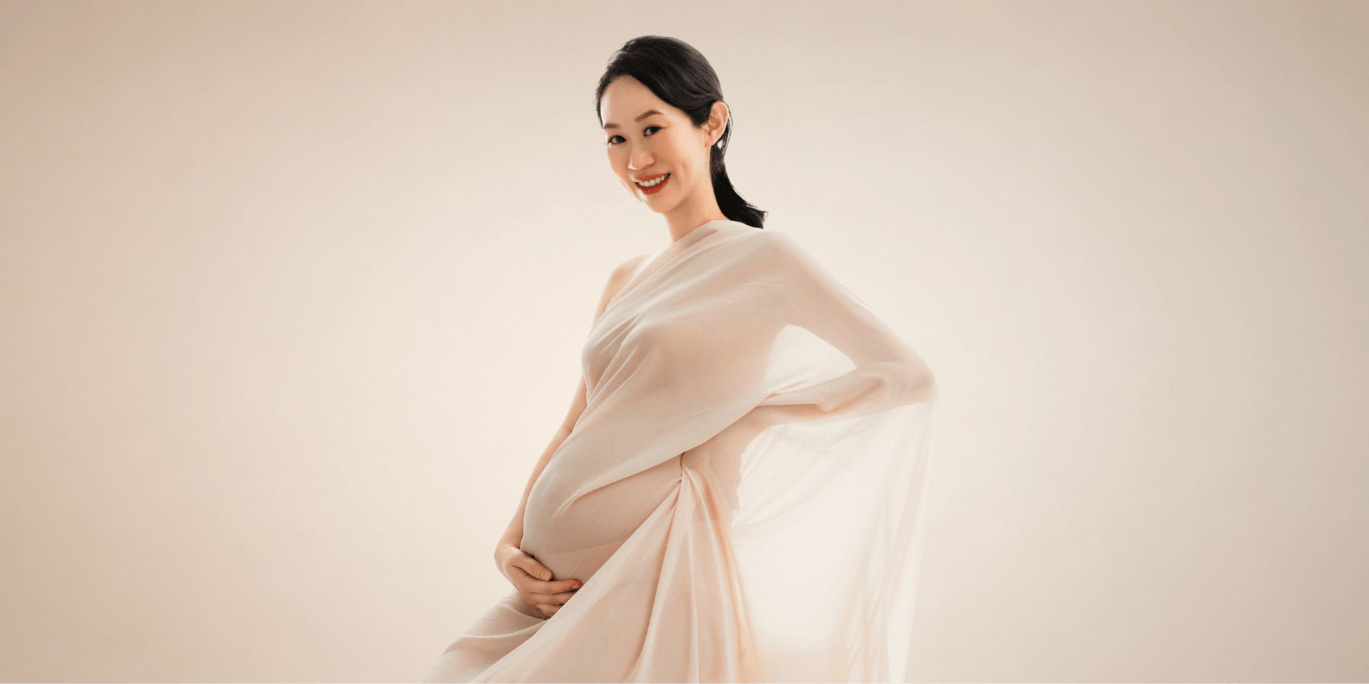 Pregnant woman in a flowing beige dress, smiling softly against a neutral background