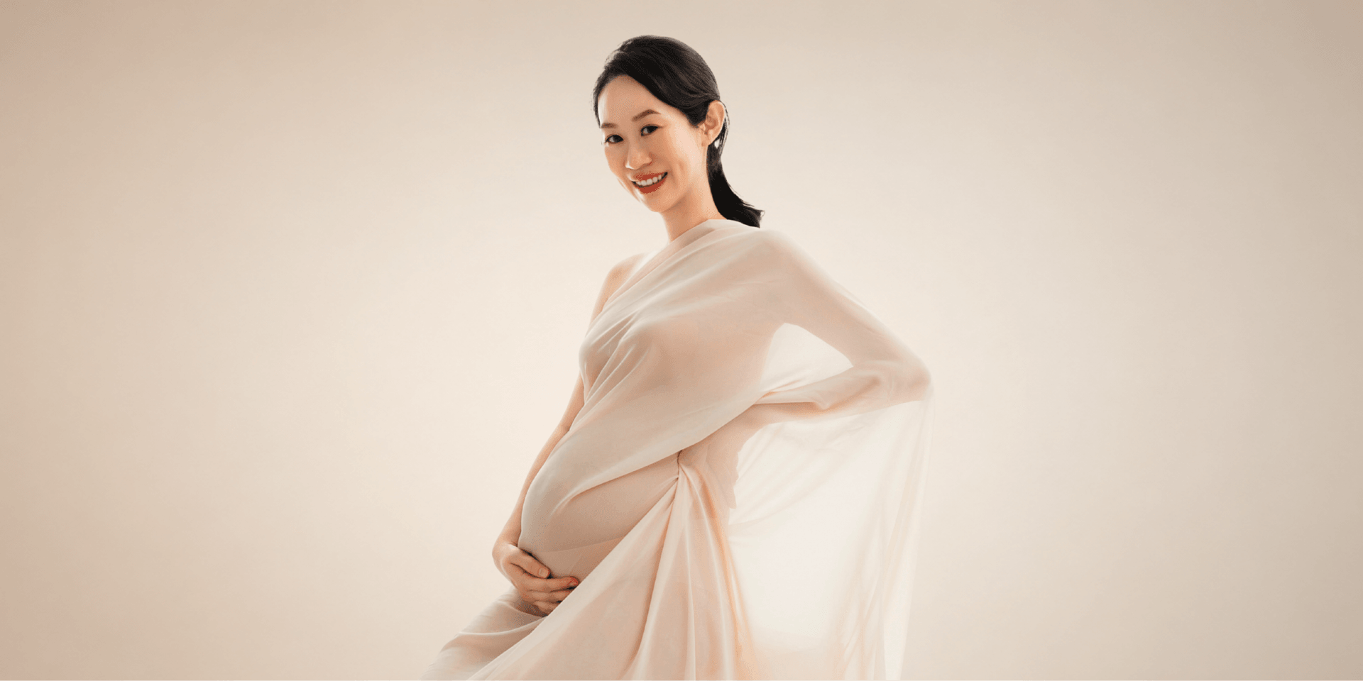 Pregnant woman in a flowing beige dress, smiling softly against a neutral background
