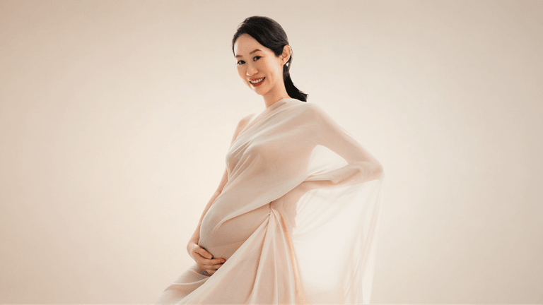 Pregnant woman in a flowing beige dress, smiling softly against a neutral background