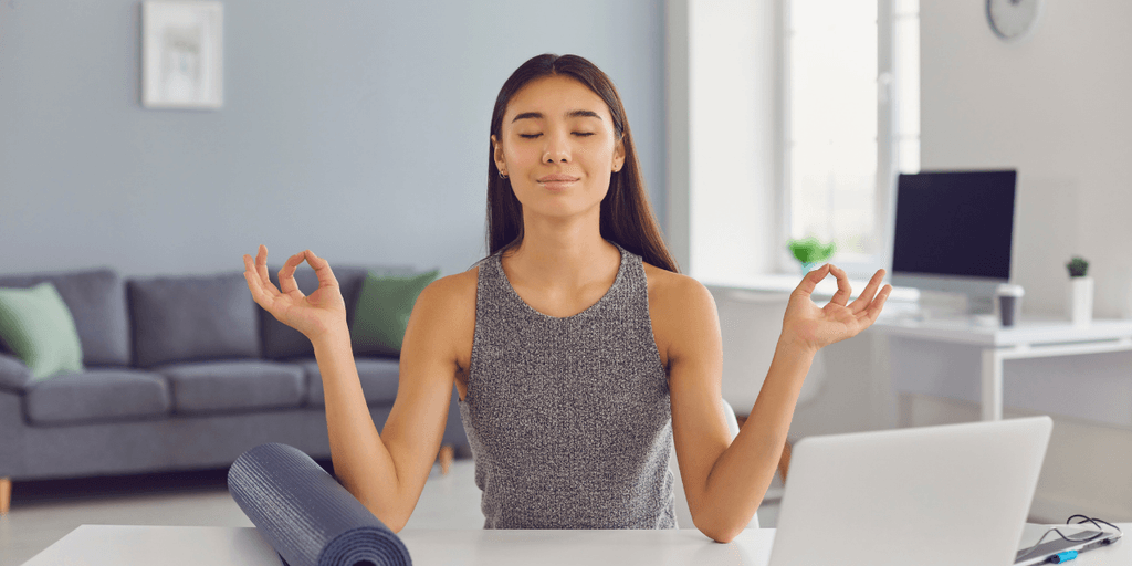 Woman meditating at a table with a yoga mat and laptop in a calm room