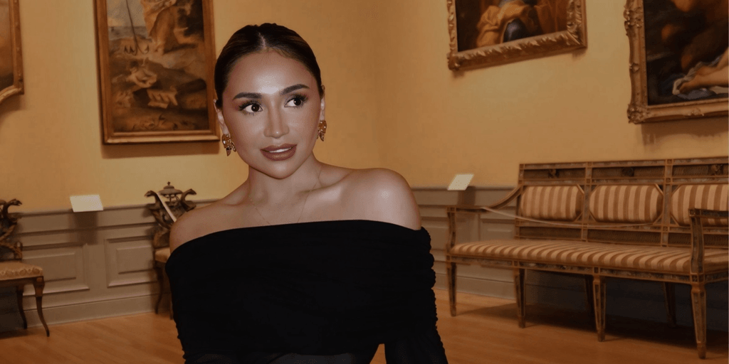 A woman in an off-shoulder black top poses in an art gallery.