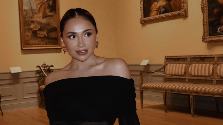 A woman in an off-shoulder black top poses in an art gallery.