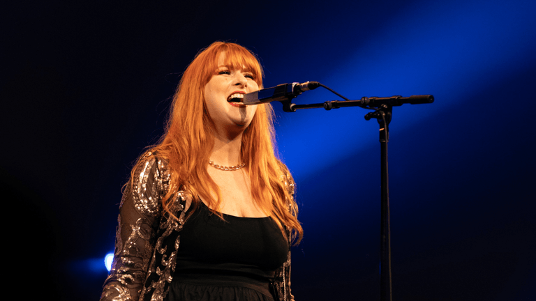 A woman with long red hair sings passionately into a microphone on stage