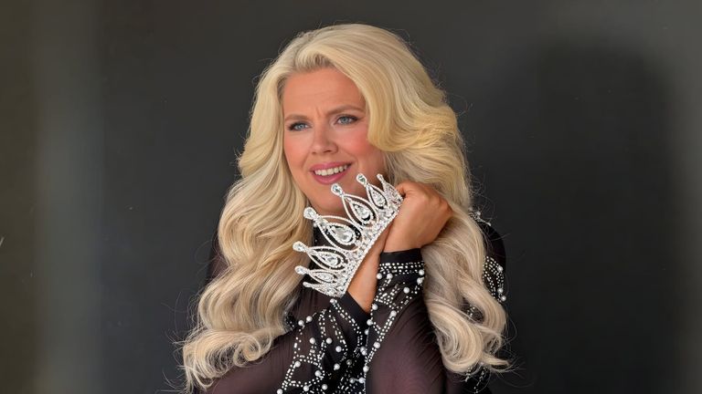 Woman with long blonde hair holding a silver tiara, wearing a sparkling black outfit, smiles warmly against a dark background.
