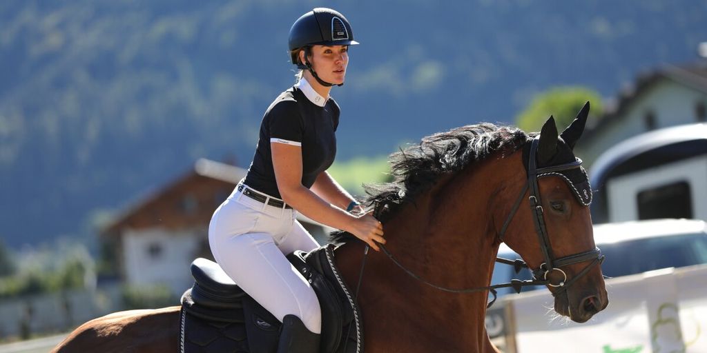 A woman in riding attire competes in showjumping, confidently riding her horse in an outdoor arena surrounded by nature.