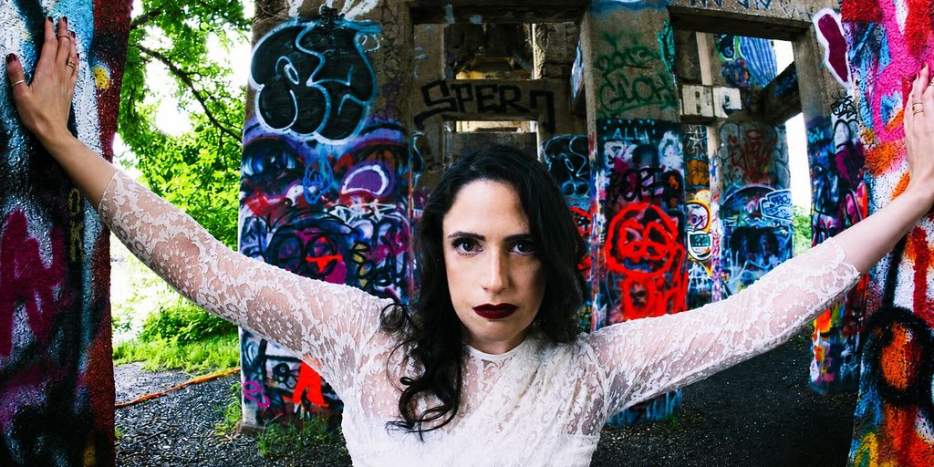 Woman in a white lace dress posing between colorful graffiti-covered concrete pillars.