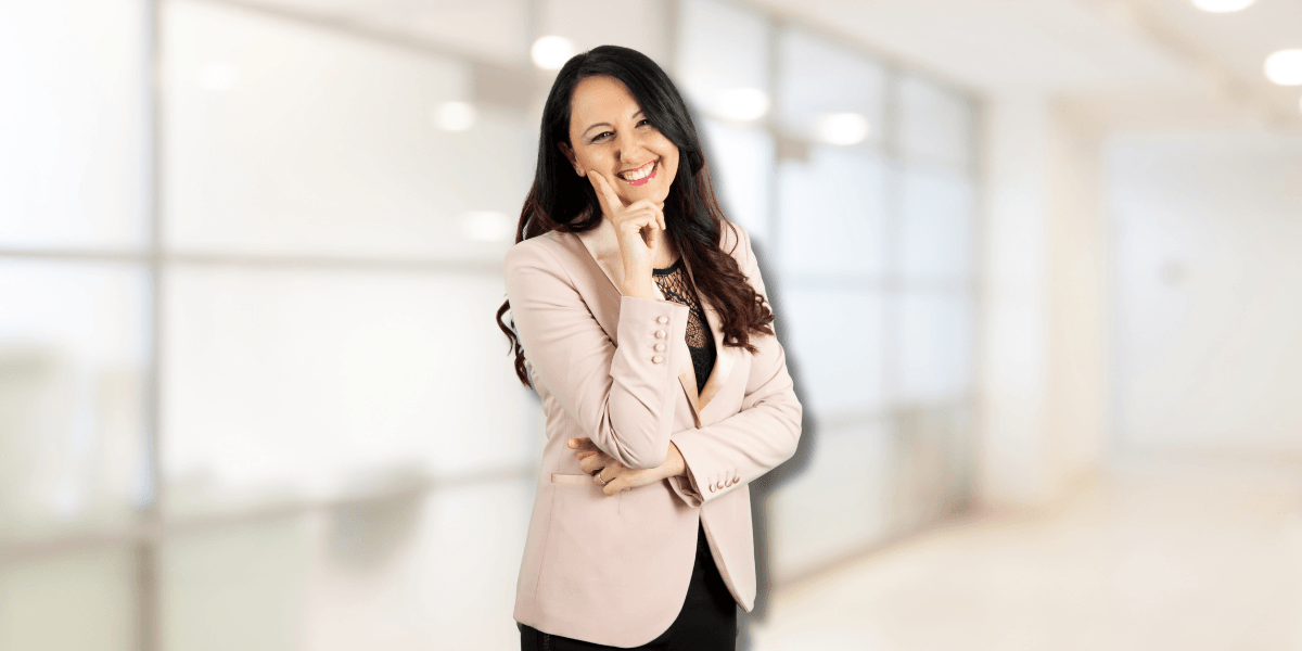 Samya Ilaria Di Donato, a confident woman in a pink blazer, smiles thoughtfully with one hand on her chin.