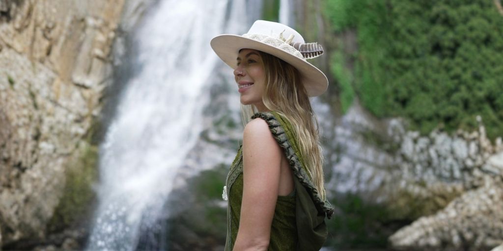 A woman stands by a waterfall, wearing a bohemian dress, wide-brimmed hat, and a serene smile, reflecting spiritual harmony.
