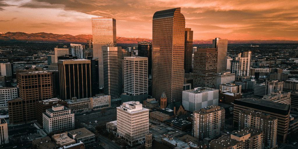 Aerial view of Denver skyline at sunset with downtown skyscrapers and Rocky Mountains in the background.