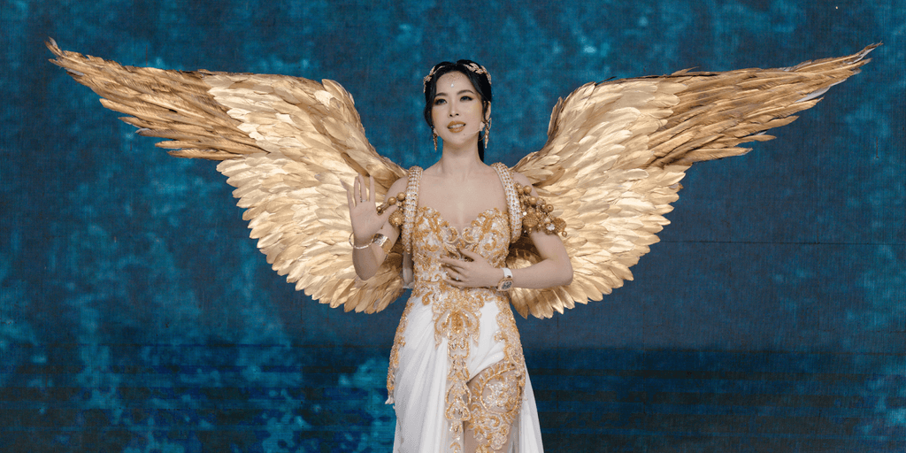 "Close-up of a model dressed in a glamorous angel-inspired costume with golden wings, giving a serene pose at a fashion show on a blue-lit stage."