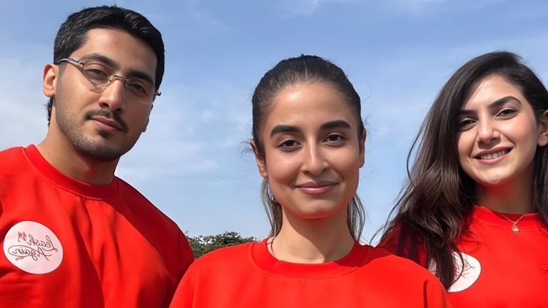 three smiling individuals wearing red LeashAgain shirts, standing together outdoors with a bright sky in the background.