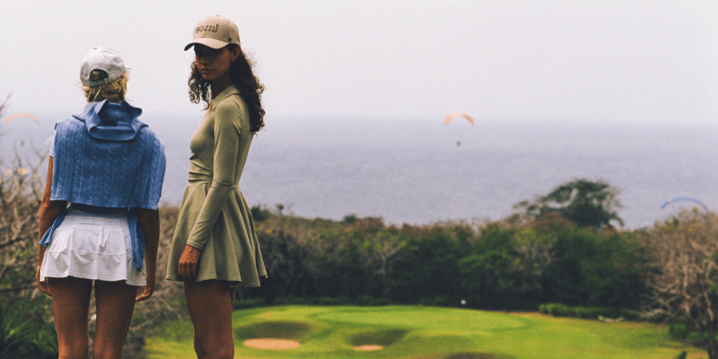 Two women in luxury headwear, overlooking a golf course with paragliders in the background, embodying elegant adventure.